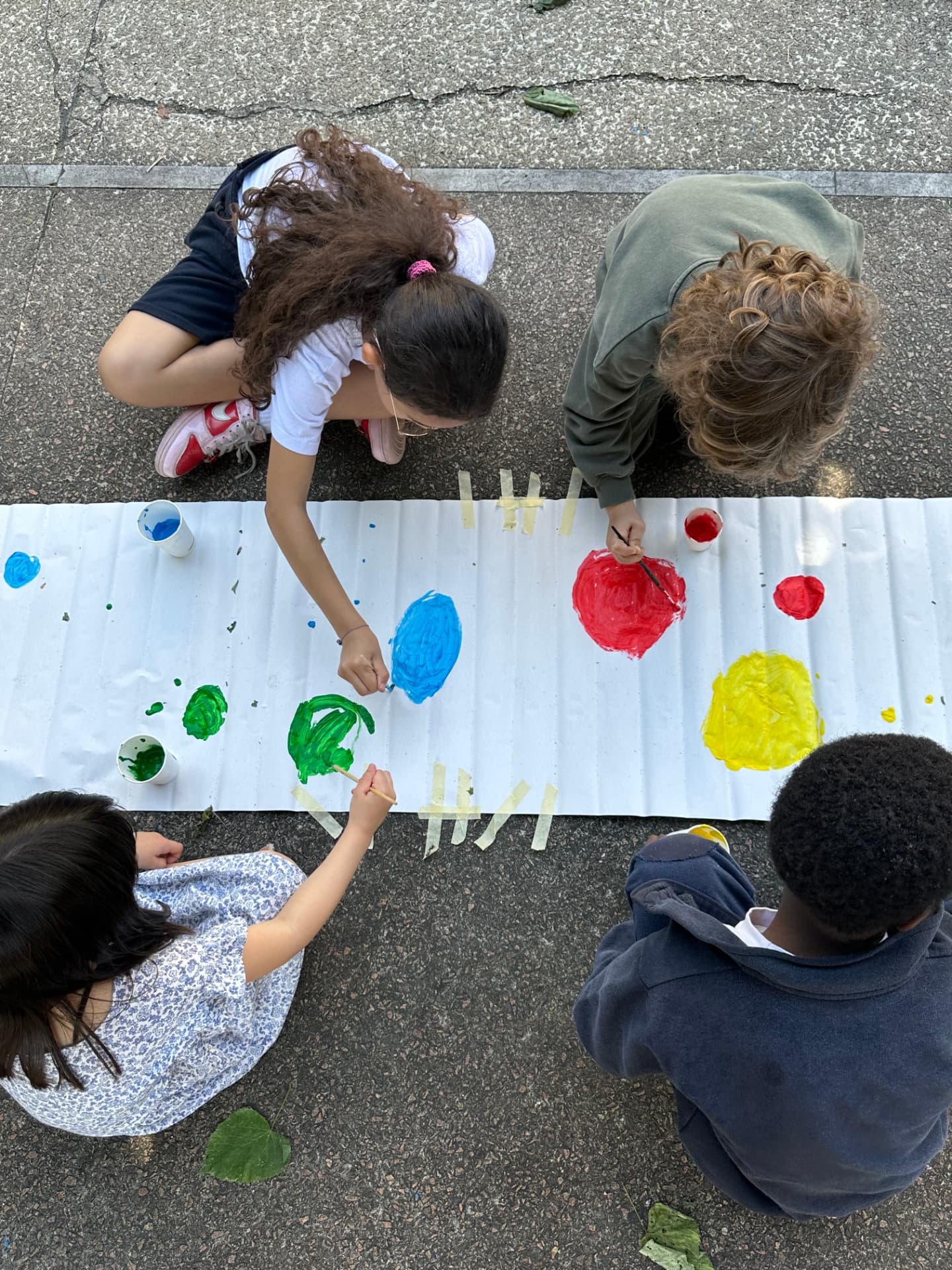 Quattro bambini dipingono insieme una lunga striscia di carta all'aperto durante un laboratorio al Buon Pastore